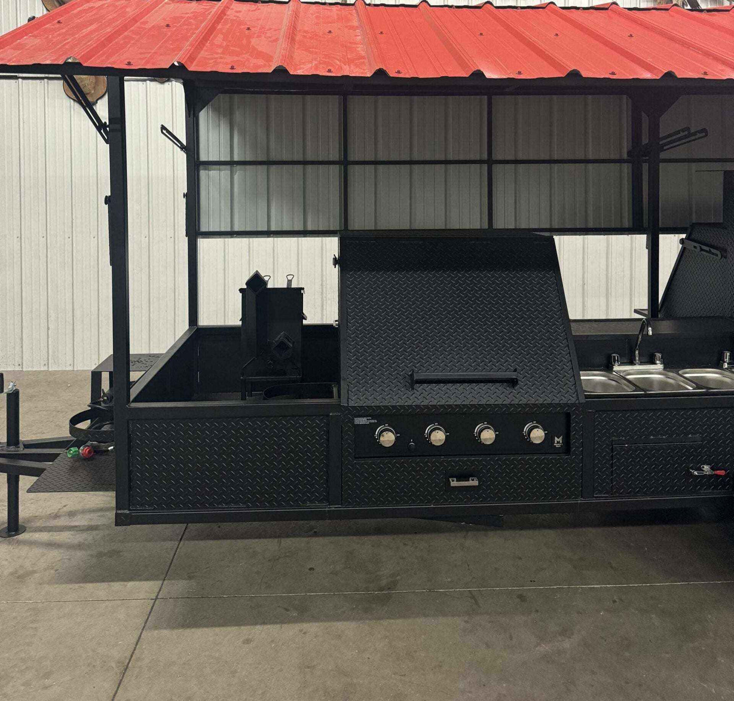 Black custom smoker on trailer with red roof, multiple cooking compartments, sink package, and propane griddle.