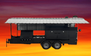 Custom Ultimate Smokehouse 72 commercial smoker with griddle, sink package, double-walled firebox, and multiple cooking racks against sunset background