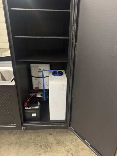 Interior storage cabinet of SNS Hospitality BBQ Trailer showing battery, water system components, and shelves