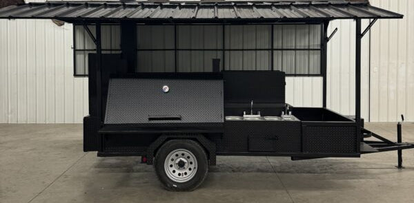 A black metal trailer-style smoker or grill stand with an open side canopy, parked indoors on a concrete floor.