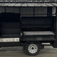 A black outdoor kitchen trailer with a corrugated metal roof and side prep table.