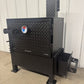 Black metal barbecue smoker with a visible brand logo against a white wall.