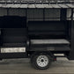 A black outdoor kitchen trailer with a corrugated metal roof and side prep table, parked on a concrete floor in front of a white wall.