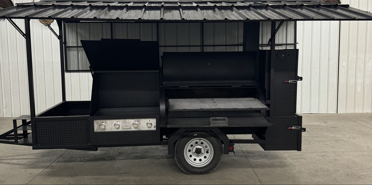 A black outdoor kitchen trailer with a corrugated metal roof and side prep table, parked on a concrete floor in front of a white wall.