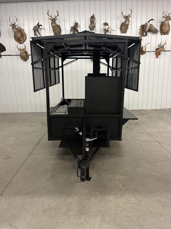 black SNS 60 smoker cabinet and grill combo trailer with fold-down roof and mounted fish fryer in indoor setting with mounted deer heads on white wall background