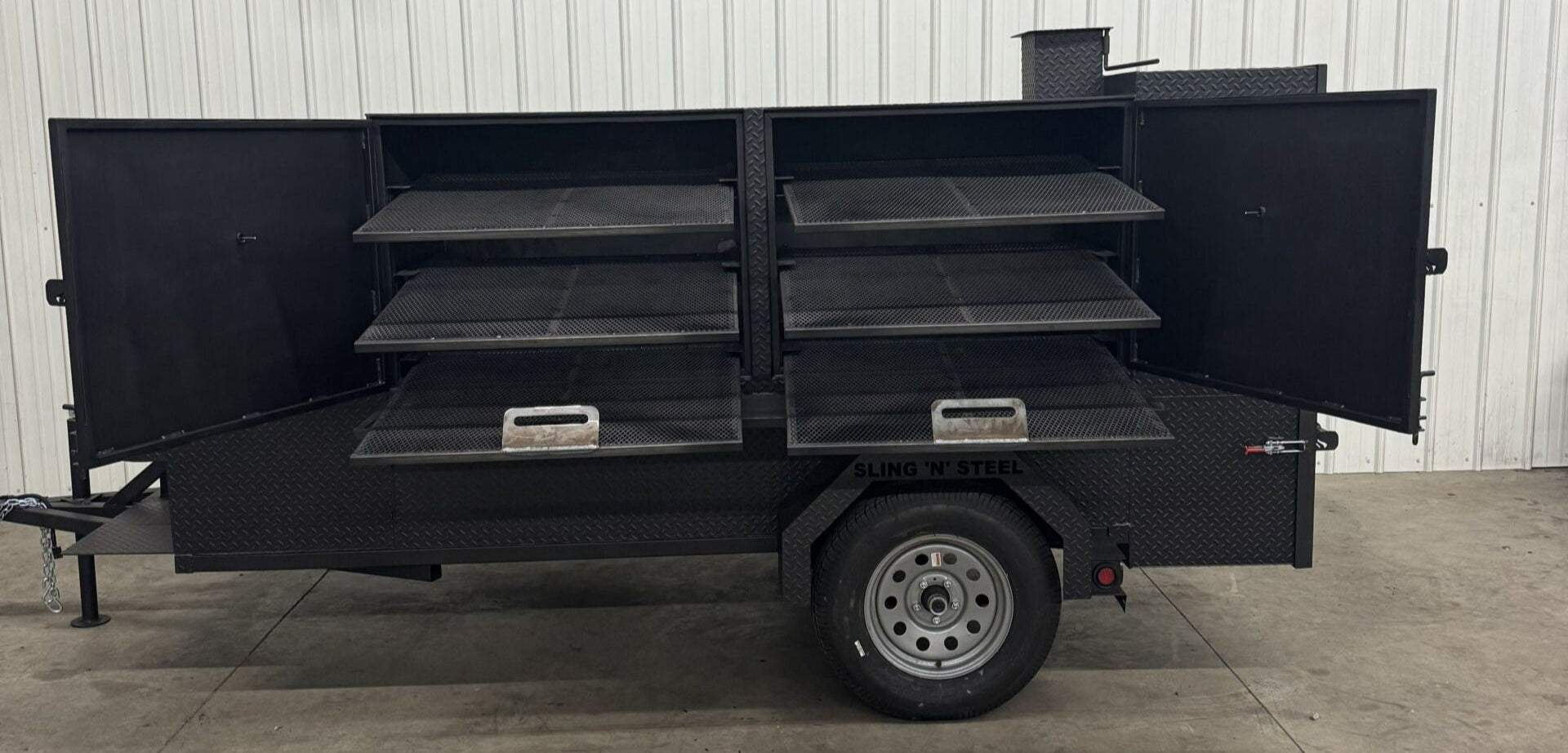 Large black SNS Beast Cabinet wood smoker with full length storage, open doors revealing multiple slide-out cooking racks and diamond tread steel finish on trailer-mounted base.