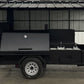 A black metal trailer-style smoker or grill stand with an open side canopy, parked indoors on a concrete floor.